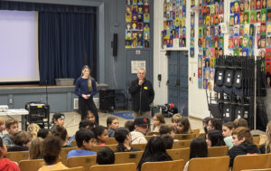 School assembly with speaker and audience seated.